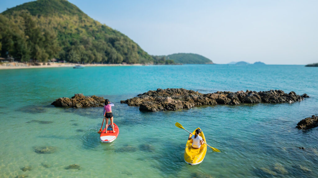 Asian athletic woman on paddle board with friend on kayak at the sea. Outdoor water sport and travel on summer holiday Thailand.